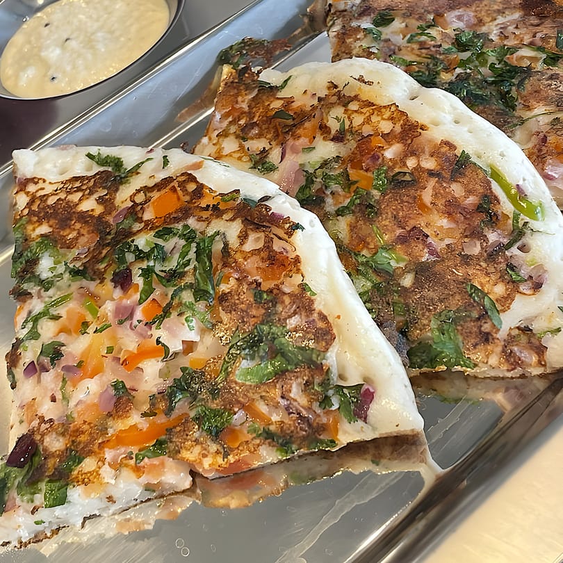 Three pieces of Indian uttapam, topped with vegetables and herbs, on a silver tray with a dipping sauce.