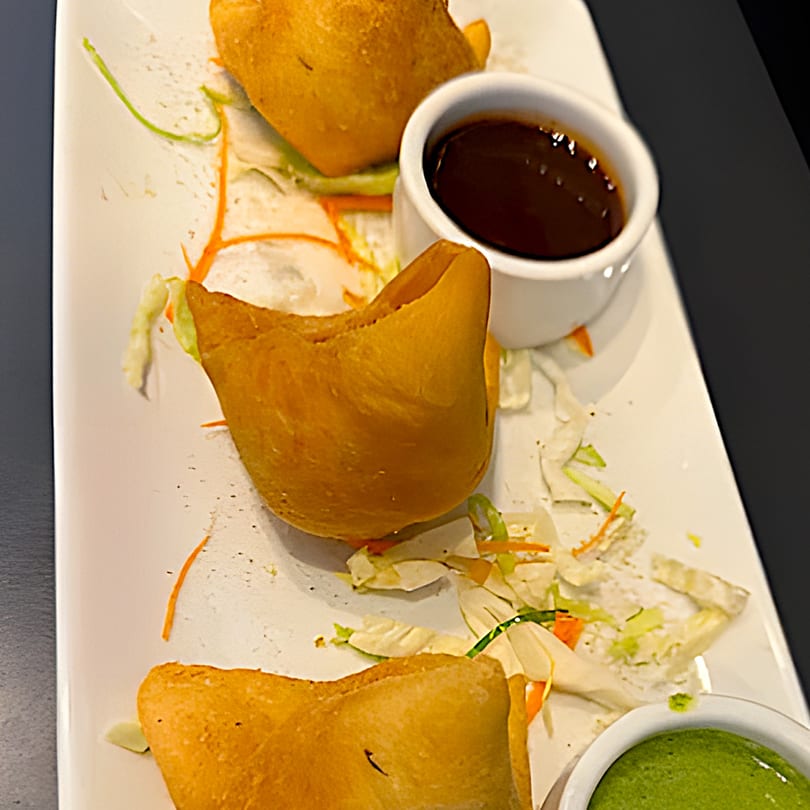 Three golden-brown samosas on a white plate with shredded vegetables and two dipping sauces.