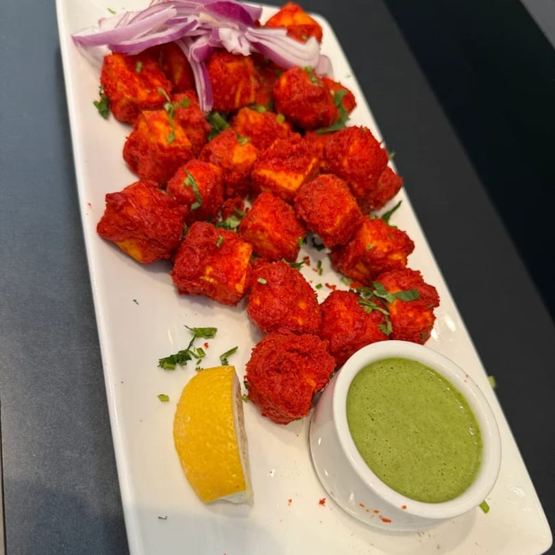 A white platter of red paneer tikka cubes, topped with red onions, a lemon wedge, and green chutney.