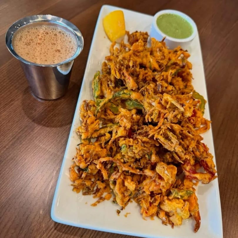 A plate of crispy Indian pakora with green chutney, a lemon wedge, and a steel cup of chai.